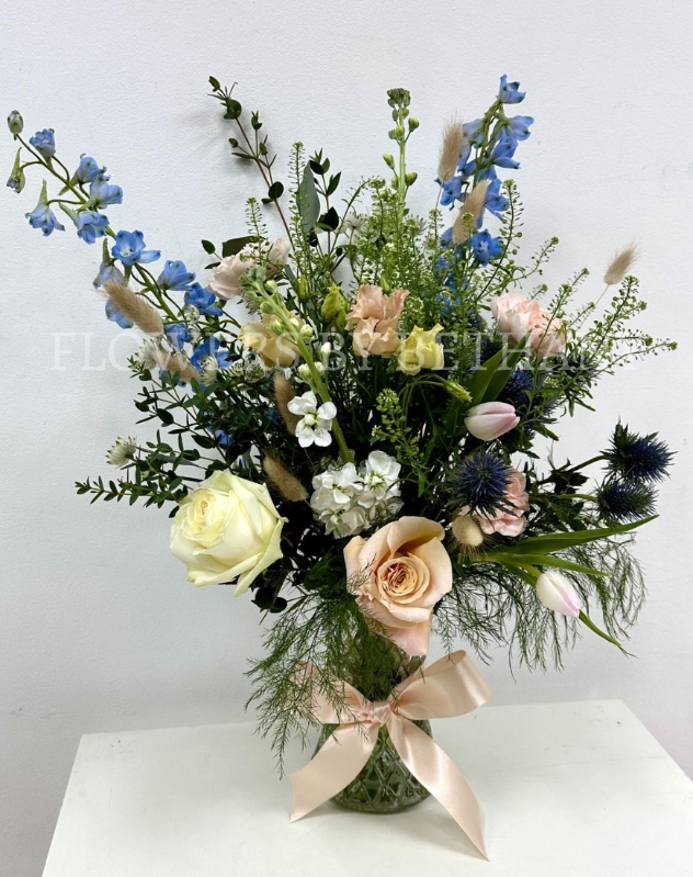 A hand tied bouquet filled with seasonal varities in peach, pink and blue tones presented in a glass vase with a bow. 