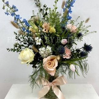 A hand tied bouquet filled with seasonal varities in peach, pink and blue tones presented in a glass vase with a bow. 
